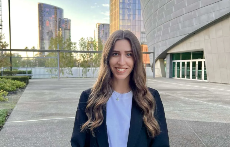 Woman with long brown hair standing in front of buildings wearing white shirt and black jacket.